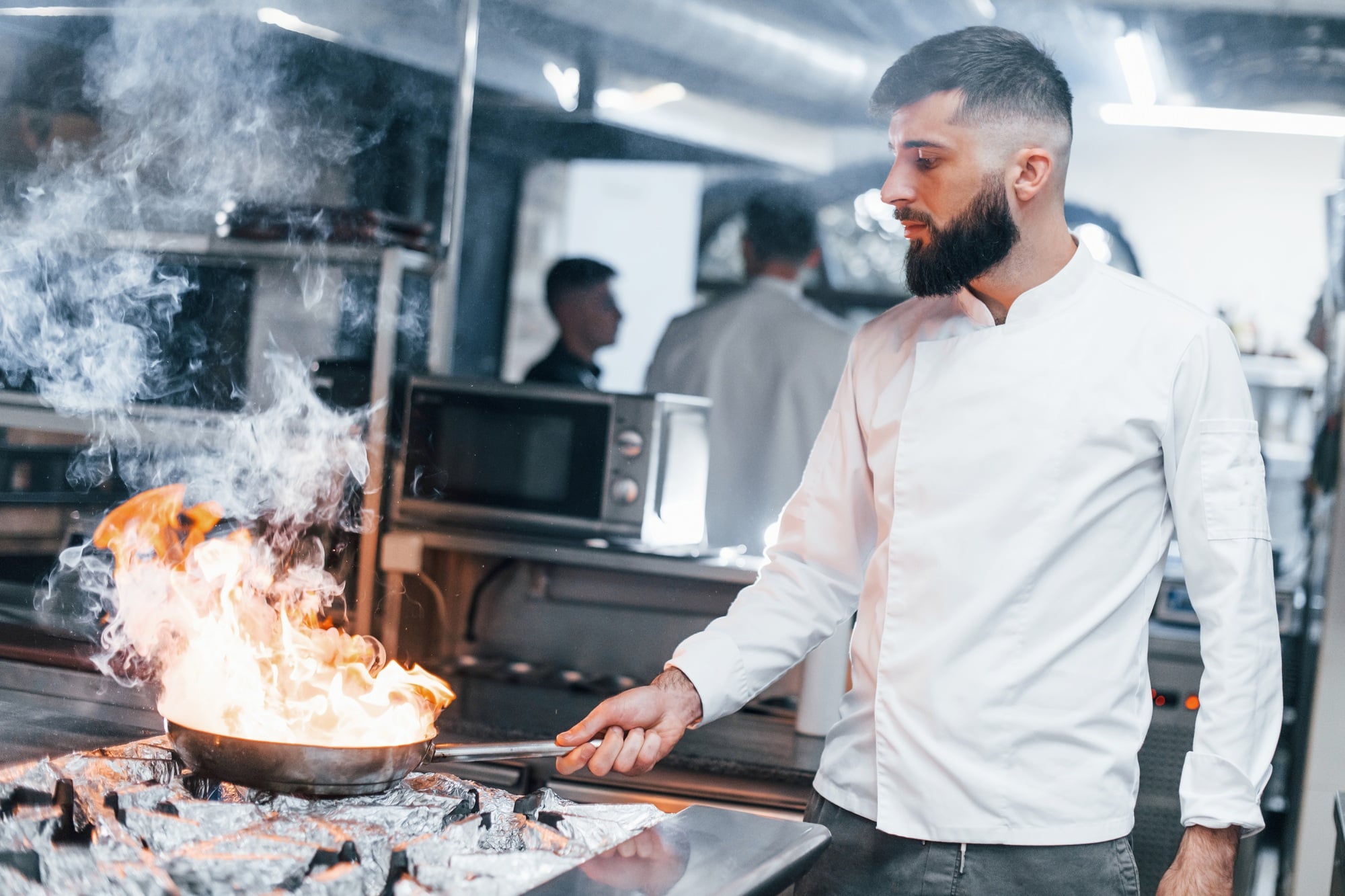 Frying pan is on fire. Chef in white uniform cooking food at kitchen. Busy day at work